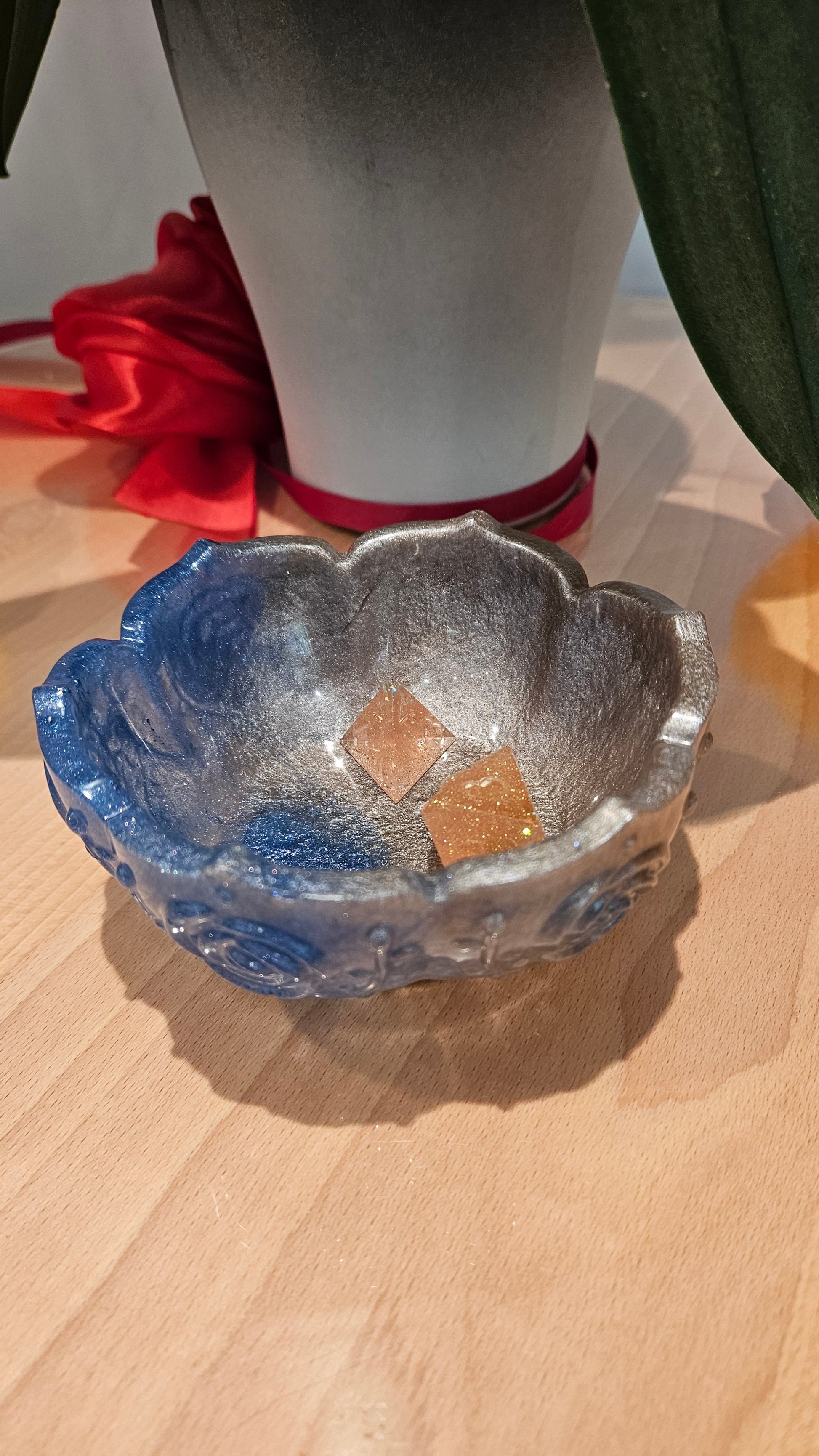 Decorative blue and silver flower-shaped  resin bowl containing dice. On a wooden table. Orchids in the background.