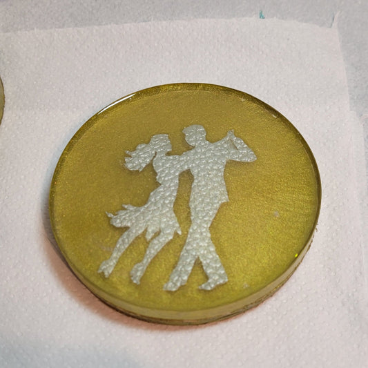 Gold coloured resin circular drinks coaster. Inside is a faux leather pair of Latin American dancers.
